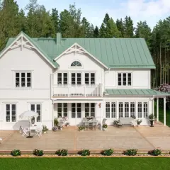 Vitt hus med torn och grönt tak, omgivet av skog och blommande syrenbuskar. En stor altan med utemöbler inbjuder till avkoppling.