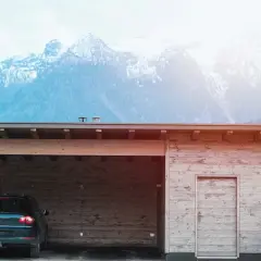 Nybyggd carport mot befintligt garage, med en bil parkerad under taket. Majestätiska berg i bakgrunden.