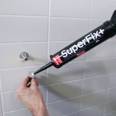 En hand använder en fogpistol med "SuperFix+" för att limma kakel. En metallbricka med ett fäste är redo att monteras.