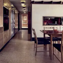 Ett kök med träinredning och ett mönstrat golv från 60-talet. Ett matsalsbord med stolar står framför en vägg med en inbyggd hylla.