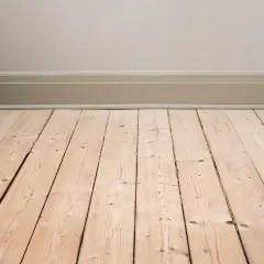 Ett ljust, obehandlat trägolv med synliga springor och kvistar. Golvet möter en ljusbeige vägg med en grågrön golvsockel.
