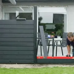 Man bygga vindskydd med vattenpass. Mörka träpaneler monteras på en altan.