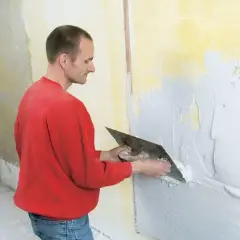 Man i röd tröja putsar en murstock med spackel och spackelspade.