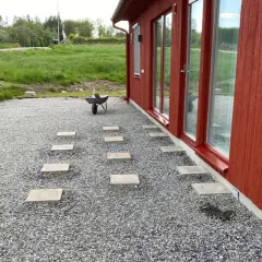 Stigande plattor leder från ett rött hus över grus. En skottkärra står redo för mer arbete med altan plintar eller plattor.