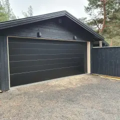 Ny, svart garageport monterad på ett mörkt trähus. En mörk trästaket står bredvid.