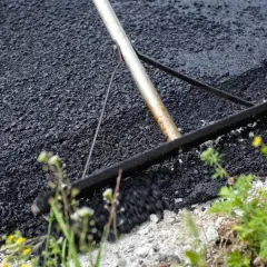 Verktyg sprider ny asfalt för att asfaltera uppfart. Grönska och gula blommor växer vid kanten.