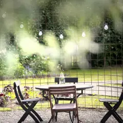 En uteplats med bord och stolar, omgiven av fina blommor och grönska. En ljusslinga med glödlampor hänger ovanför.