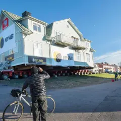 Ett stort hus är på väg att flytta. En man med cykel stannar för att titta på det imponerande arbetet.