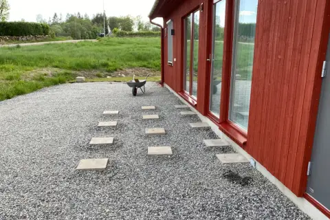 Stigande plattor leder från ett rött hus över grus. En skottkärra står redo för mer arbete med altan plintar eller plattor.