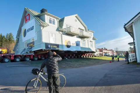 Ett stort hus är på väg att flytta. En man med cykel stannar för att titta på det imponerande arbetet.