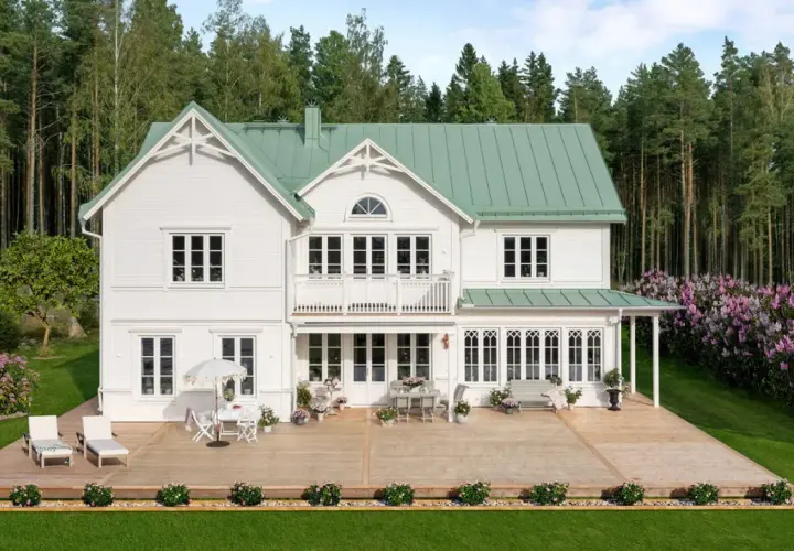 Vitt hus med torn och grönt tak, omgivet av skog och blommande syrenbuskar. En stor altan med utemöbler inbjuder till avkoppling.