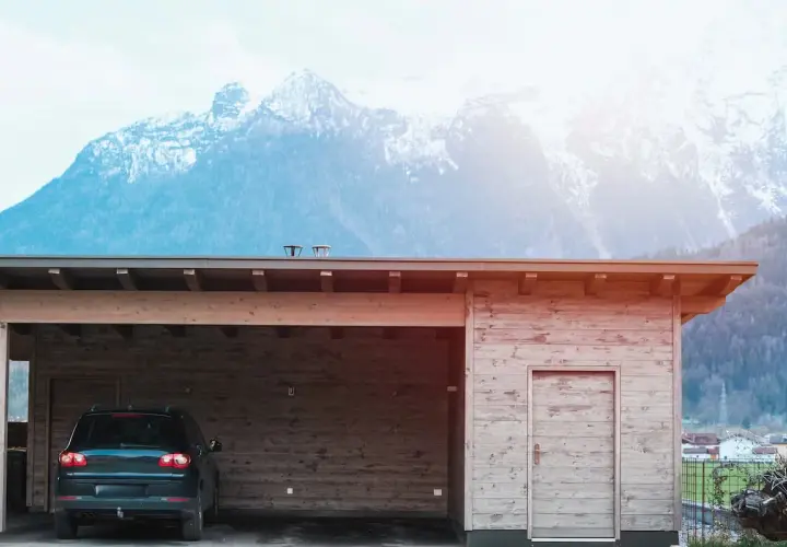 Nybyggd carport mot befintligt garage, med en bil parkerad under taket. Majestätiska berg i bakgrunden.