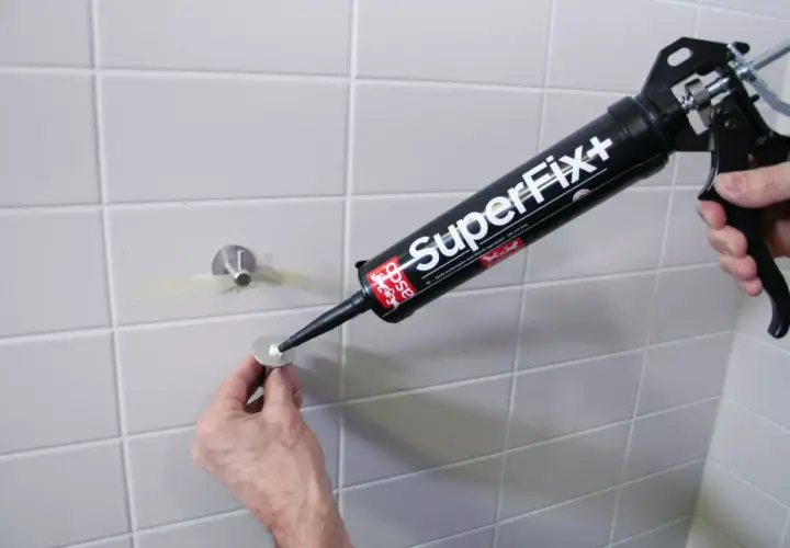 En hand använder en fogpistol med "SuperFix+" för att limma kakel. En metallbricka med ett fäste är redo att monteras.