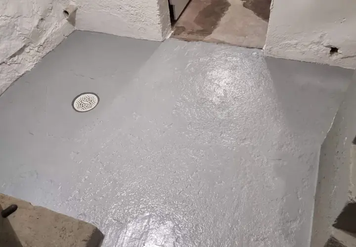 Nyligen behandlat betonggolv i källaren med en grå färg. Golvet har en avloppsbrunn och synliga trappsteg i bakgrunden.