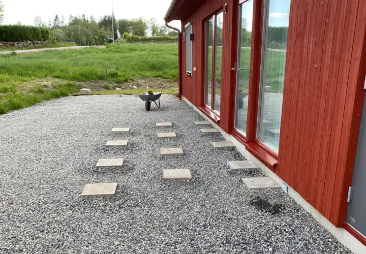 Stigande plattor leder från ett rött hus över grus. En skottkärra står redo för mer arbete med altan plintar eller plattor.