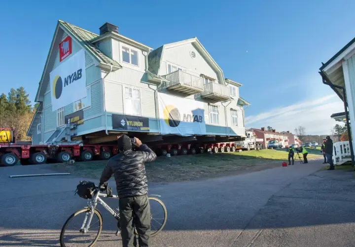 Ett stort hus är på väg att flytta. En man med cykel stannar för att titta på det imponerande arbetet.