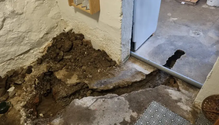 Hål i betonggolvet visar på pågående arbete med att laga rör. Jord och sten ligger uppgrävt vid väggen.