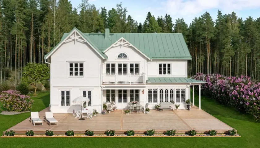 Vitt hus med torn och grönt tak, omgivet av skog och blommande syrenbuskar. En stor altan med utemöbler inbjuder till avkoppling.