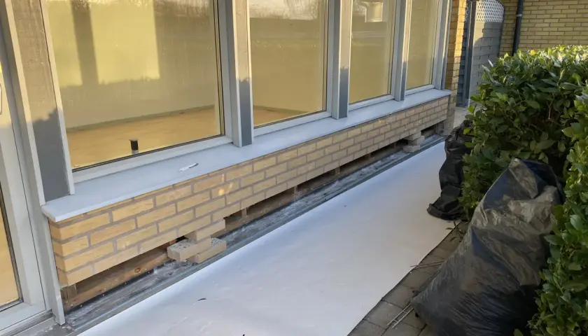 Arbete pågår för att byta syll. Nytt tegel läggs längs husgrunden, med synliga träbjälkar under.