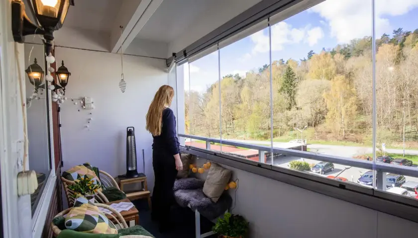 En kvinna njuter av utsikten från en mysig, inglasad balkong. Perfekt liten balkong inspiration med korgstolar och en sittbänk.