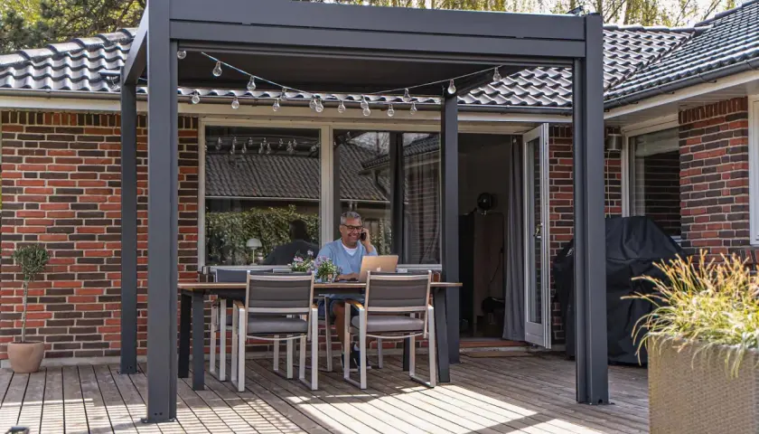 Man pratar i telefon vid ett bord under en modern pergola med vasstak. En laptop står på bordet.