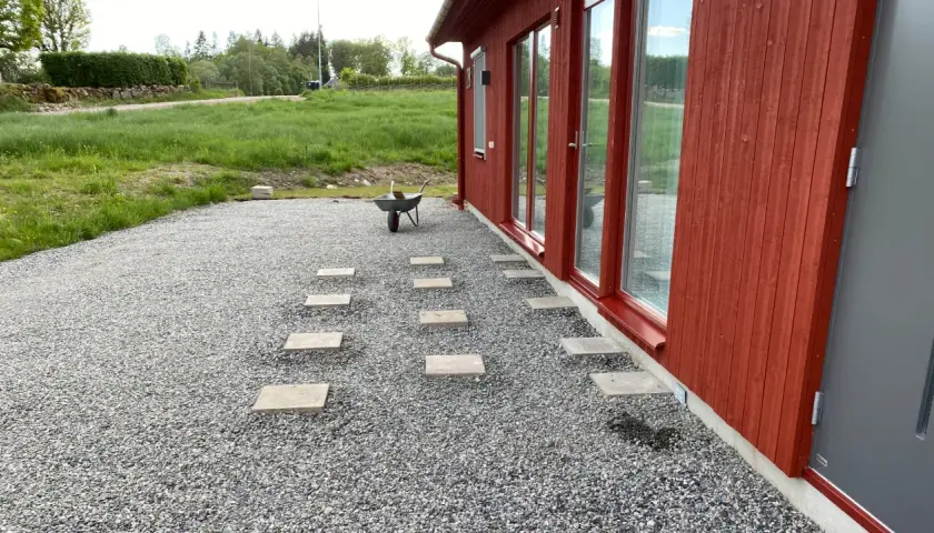 Stigande plattor leder från ett rött hus över grus. En skottkärra står redo för mer arbete med altan plintar eller plattor.