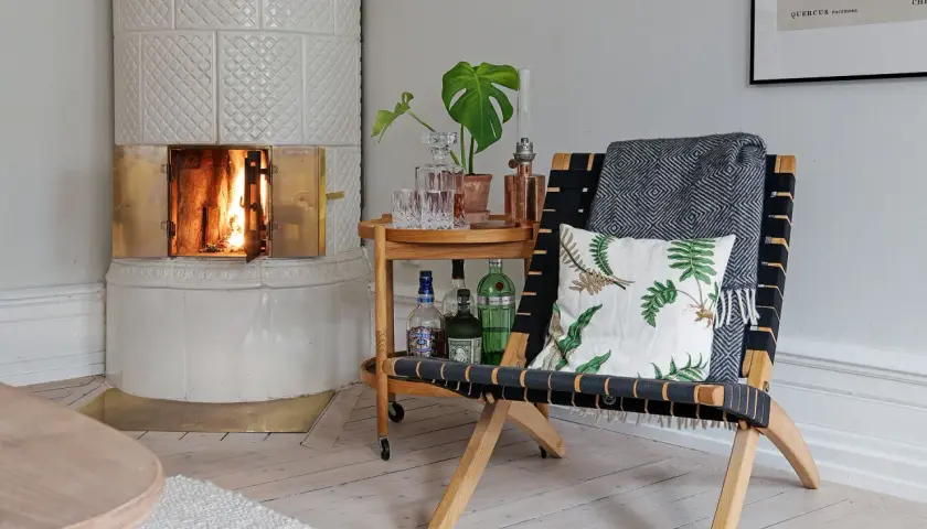 En vacker, vit kakelugn med en sprakande eld. Bredvid står en fåtölj med pläd och kudde, samt ett serveringsbord med drinkar. Perfekt för att renovera kakelugn och skapa mysfaktor.