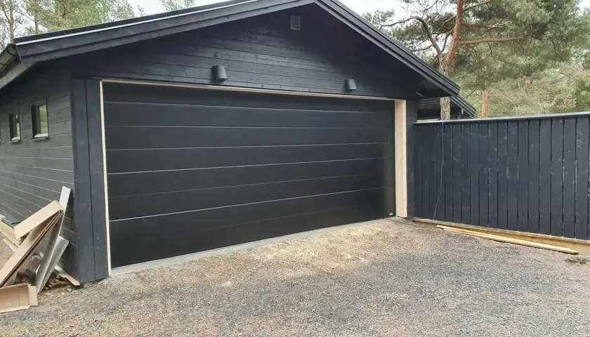 Ny, svart garageport monterad på ett mörkt trähus. En mörk trästaket står bredvid.