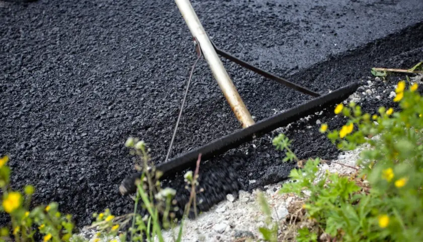 Verktyg sprider ny asfalt för att asfaltera uppfart. Grönska och gula blommor växer vid kanten.