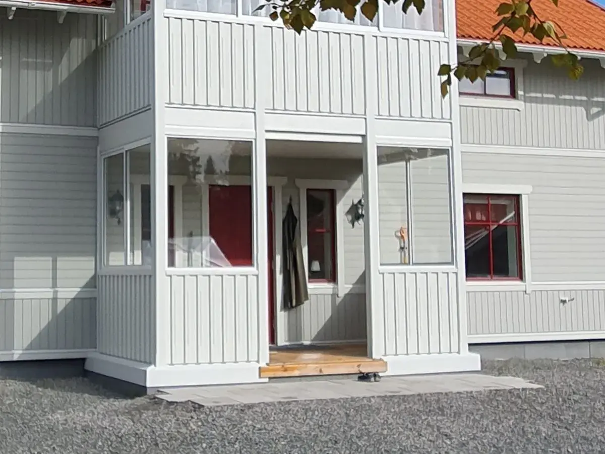 Nybyggd farstukvist med vita träpaneler och glaspartier. En röd ytterdörr syns innanför.