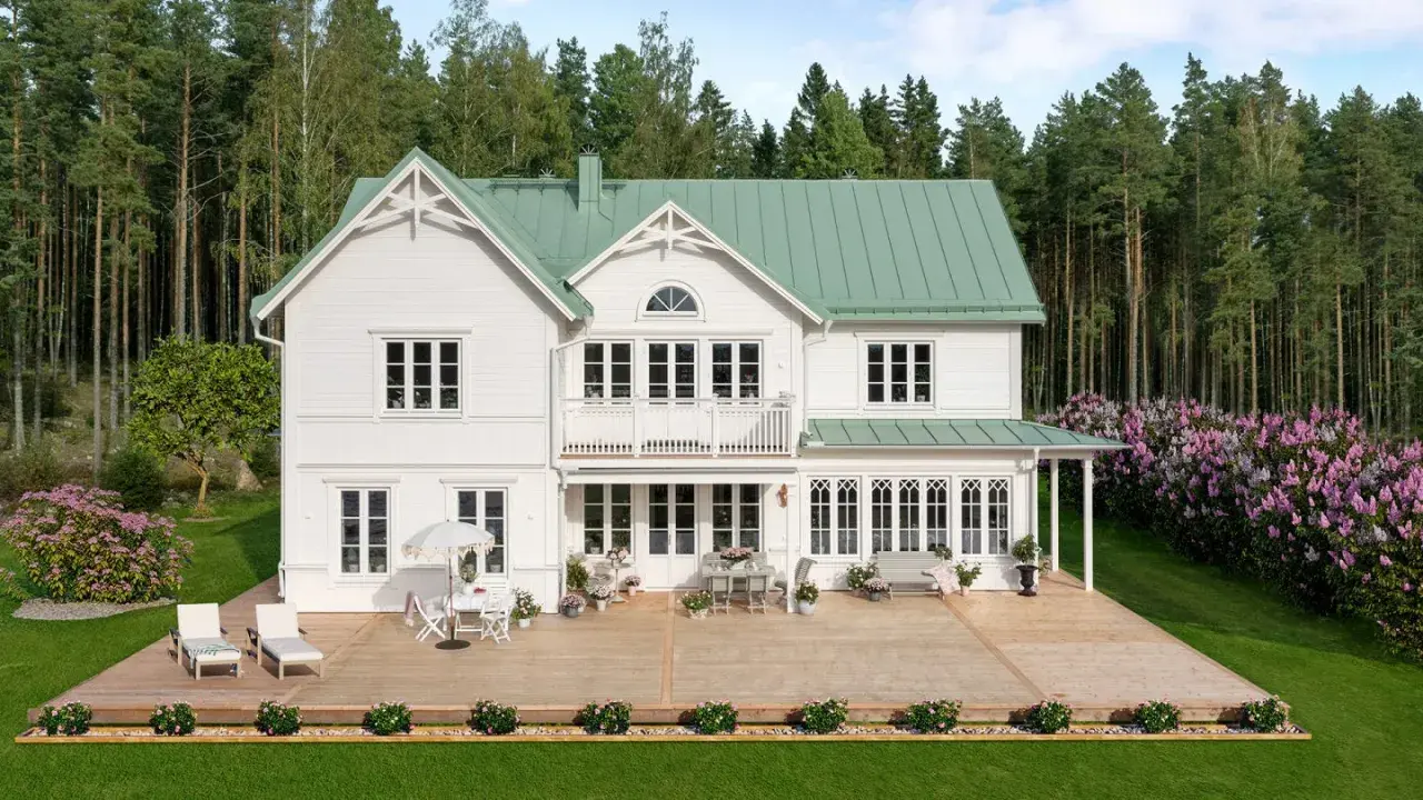 Vitt hus med torn och grönt tak, omgivet av skog och blommande syrenbuskar. En stor altan med utemöbler inbjuder till avkoppling.