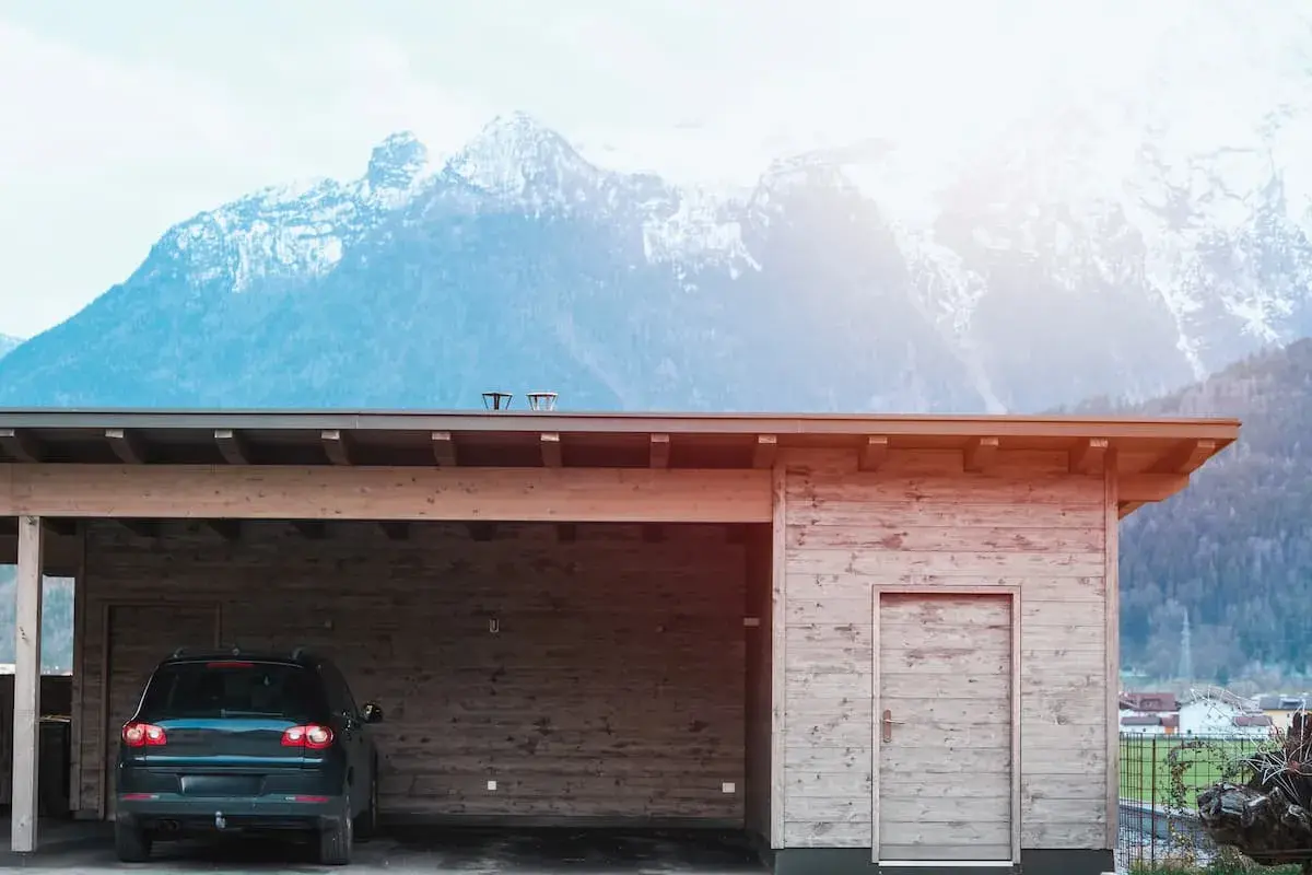 Nybyggd carport mot befintligt garage, med en bil parkerad under taket. Majestätiska berg i bakgrunden.