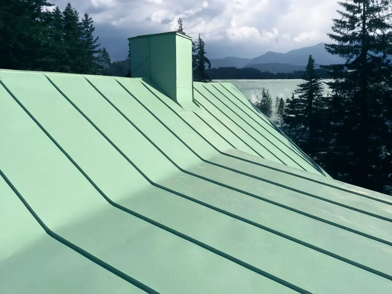 Grönt plåttak med skorsten, vacker utsikt över sjö och berg. Tänk på kostnad plåttak vid renovering.