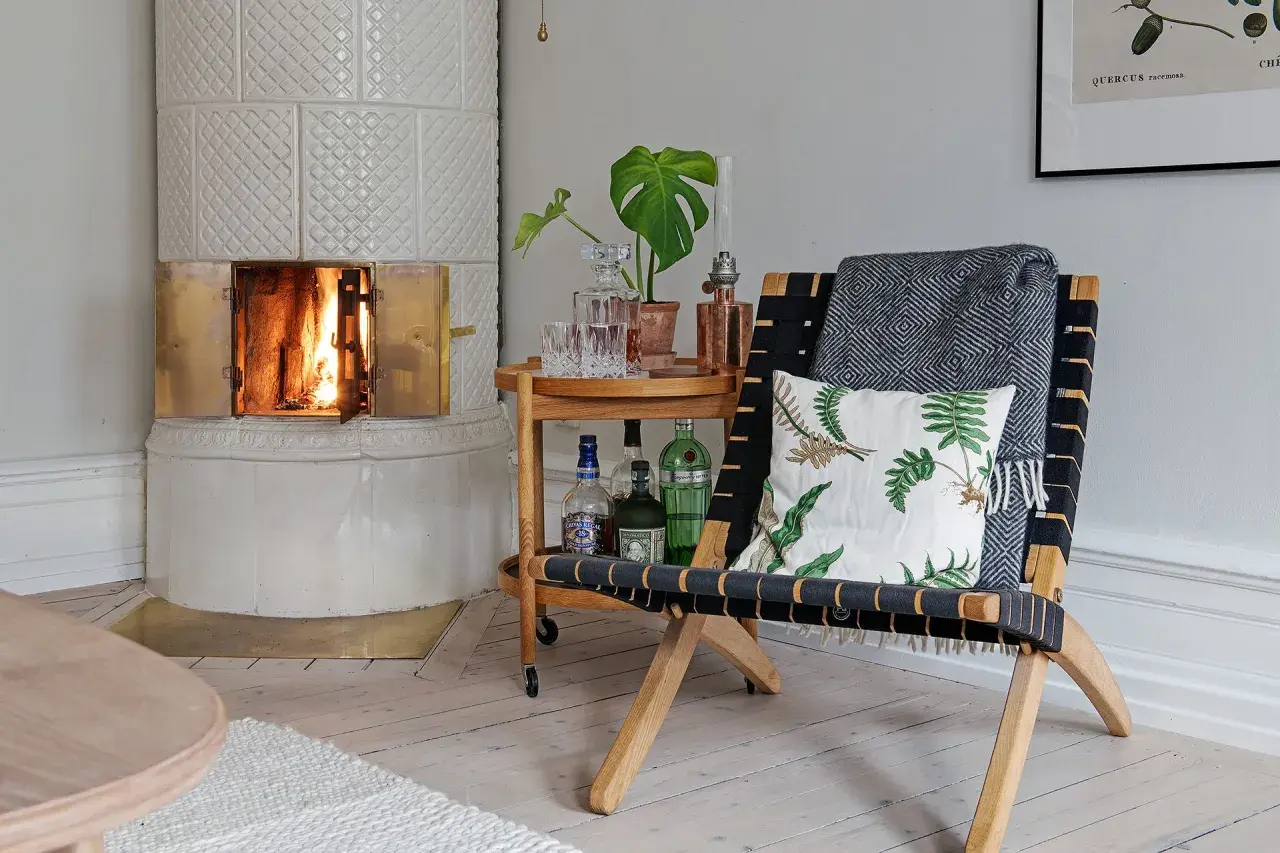 En vacker, vit kakelugn med en sprakande eld. Bredvid står en fåtölj med pläd och kudde, samt ett serveringsbord med drinkar. Perfekt för att renovera kakelugn och skapa mysfaktor.