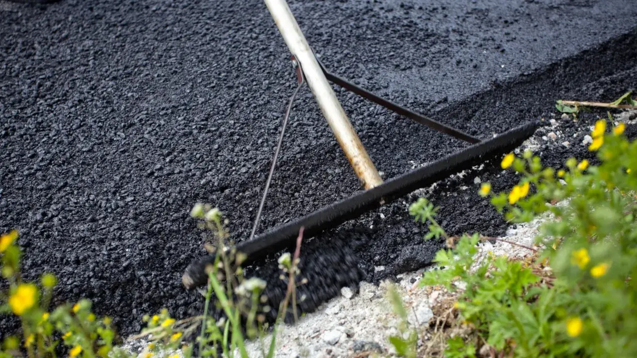 Verktyg sprider ny asfalt för att asfaltera uppfart. Grönska och gula blommor växer vid kanten.