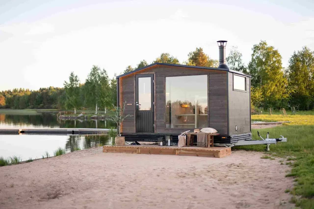 En mörk träbastu på hjul vid en sjö. Kanske krävs bygglov för denna mobila bastu?