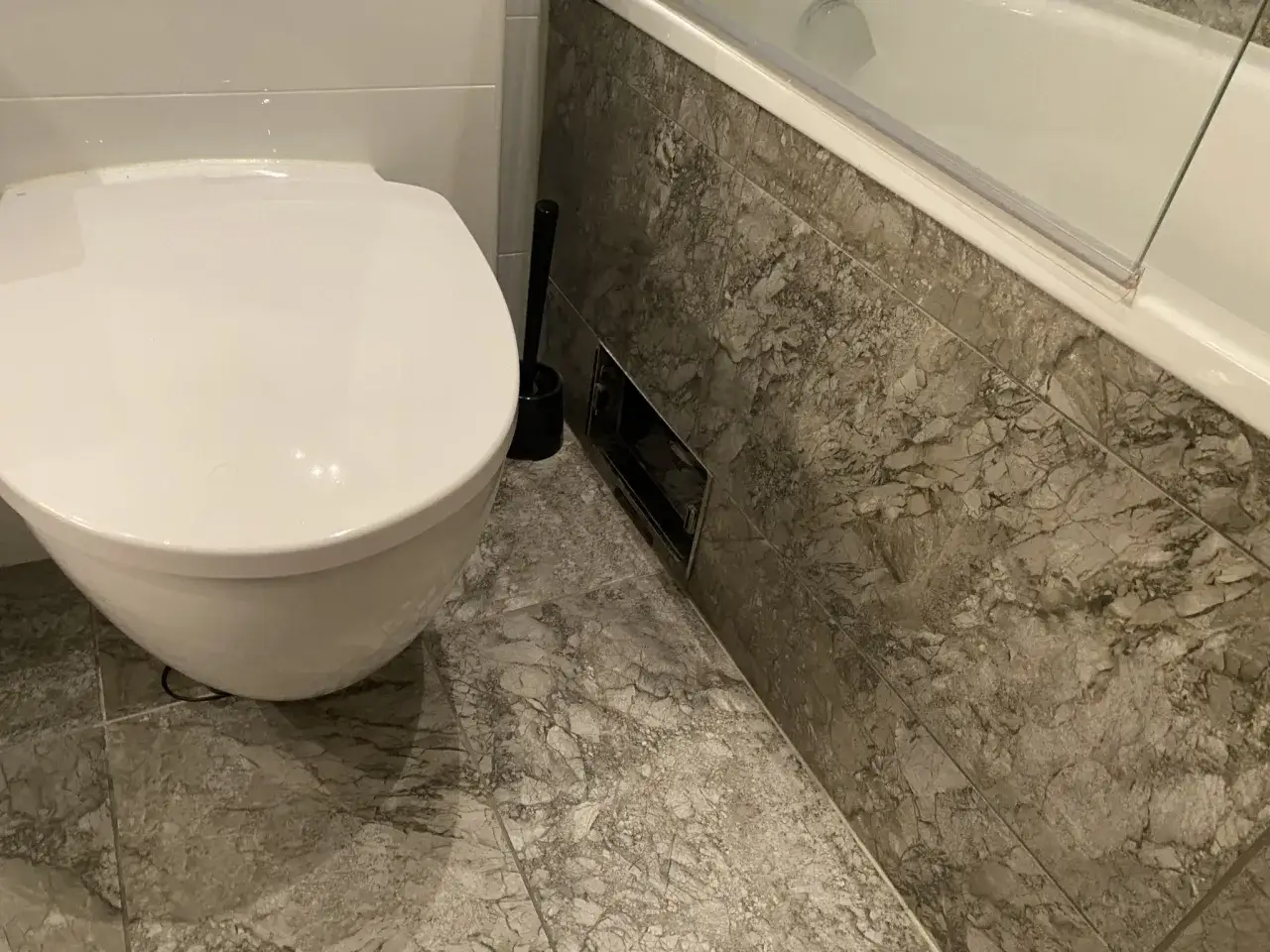 En vit toalett och ett badkar med marmorliknande kakel. En toalettborste står bredvid. Det luktar avlopp i badrummet.