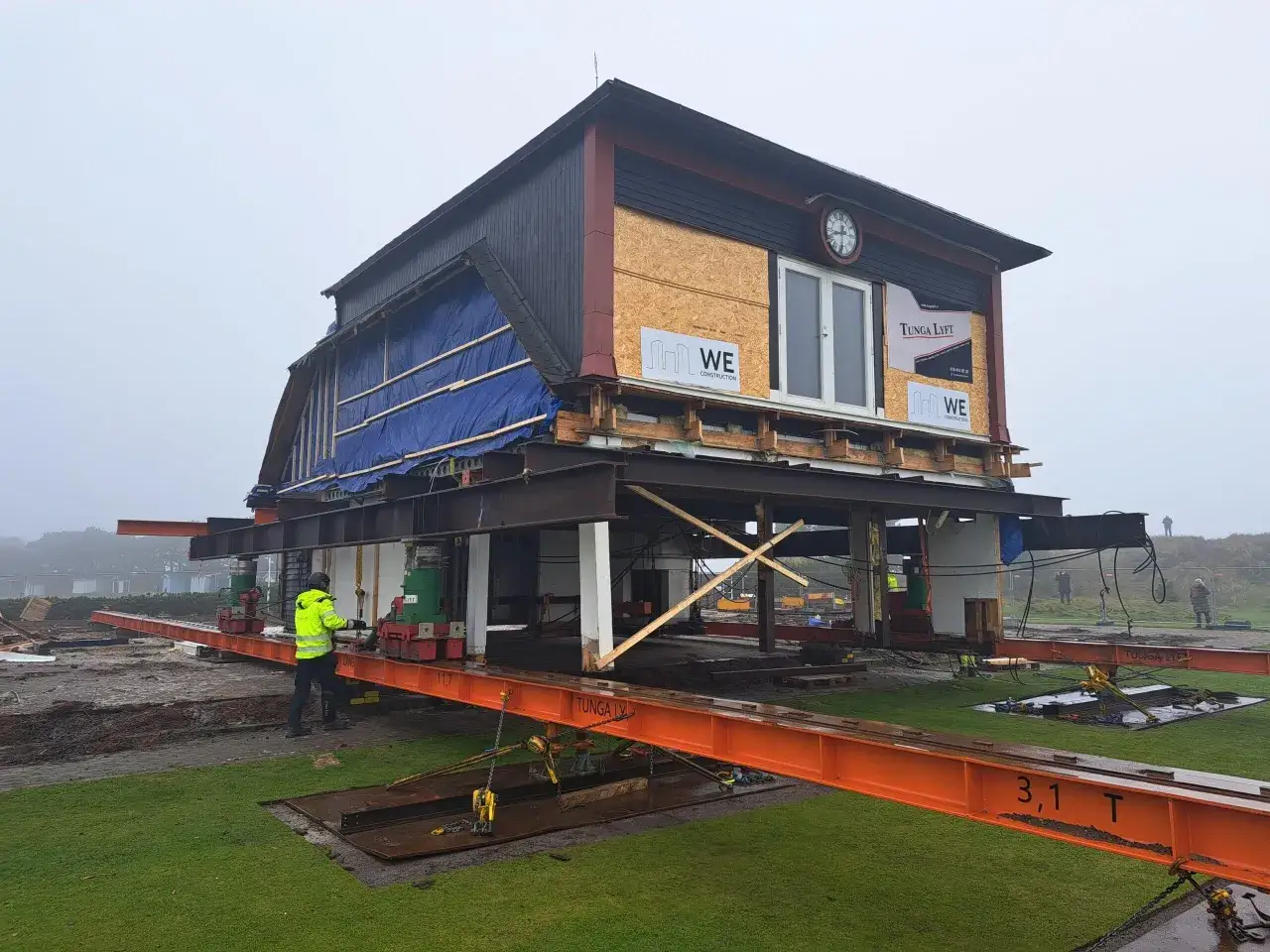 Ett hus lyfts och förbereds för att flytta. En person i varselkläder övervakar arbetet.