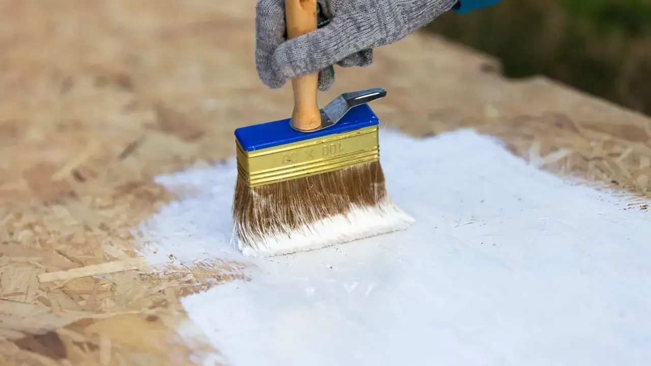 En hand med handske m&aring;lar OSB-skiva vit med en bred pensel.