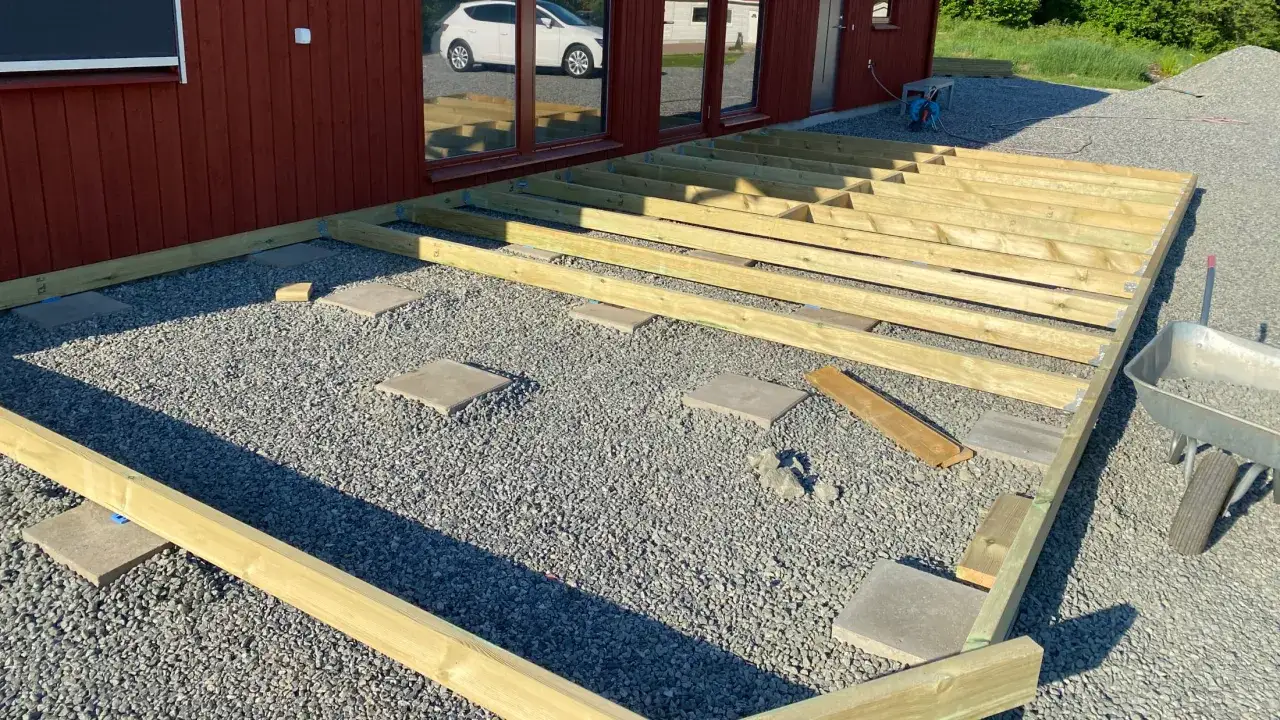 Byggnation av altan med träreglar som vilar på stenplattor. Grus och en skottkärra syns i bakgrunden.