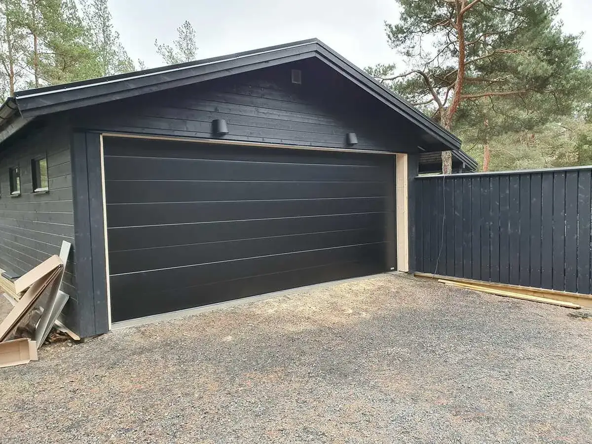 Ny, svart garageport installerad p&aring; ett m&ouml;rkt tr&auml;hus. En m&ouml;rk tr&auml;staket st&aring;r bredvid.