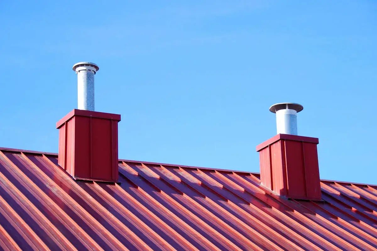R&ouml;tt pl&aring;ttak med tv&aring; r&ouml;da skorstenar och metallr&ouml;r mot bl&aring; himmel. En bra investering f&ouml;r att f&aring; koll p&aring; kostnad pl&aring;ttak.