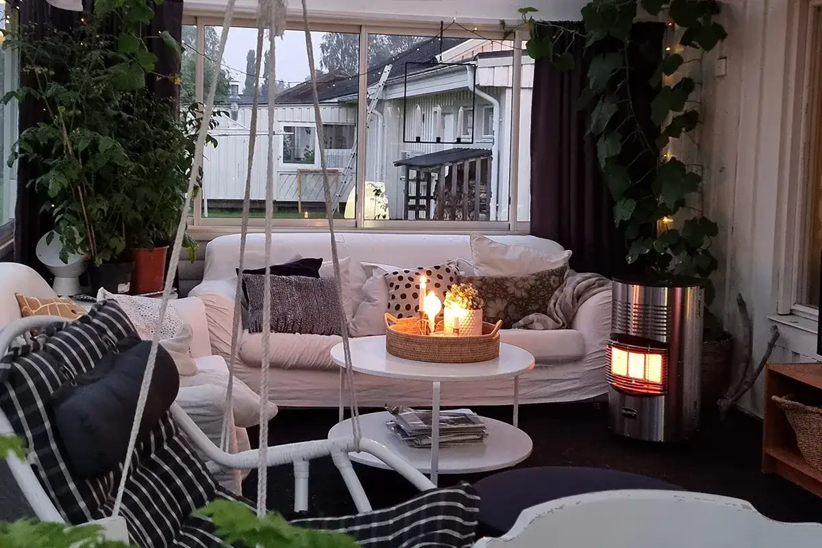 Ett mysigt uterum med soffa, kuddar, ljus och en v&auml;rmare. Utanf&ouml;r syns hus och tr&auml;dg&aring;rd.