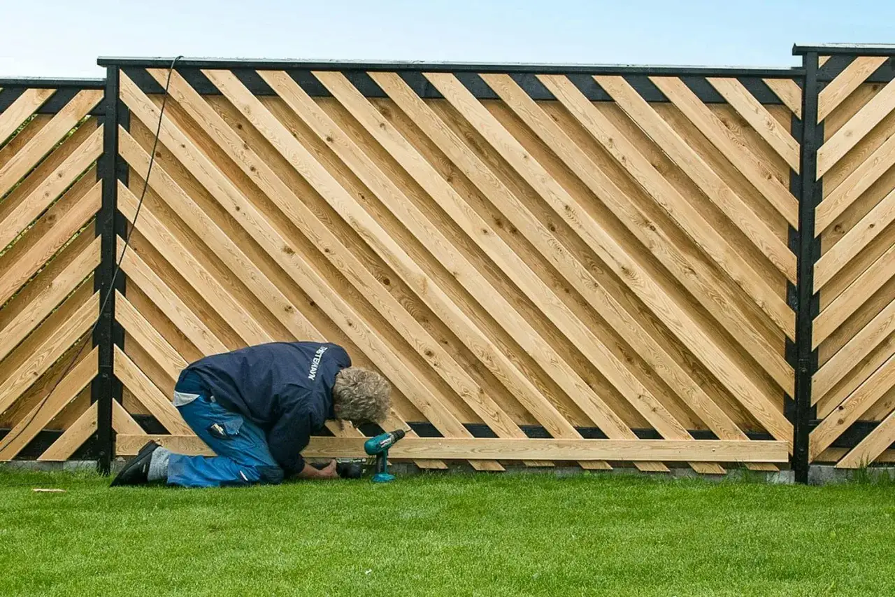 Man bygger plank med snedställda träribbor och svarta stolpar. Gräsmatta och blå himmel syns.