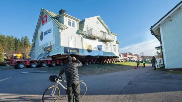 Ett stort hus är på väg att flytta. En man med cykel stannar för att titta på det imponerande arbetet.
